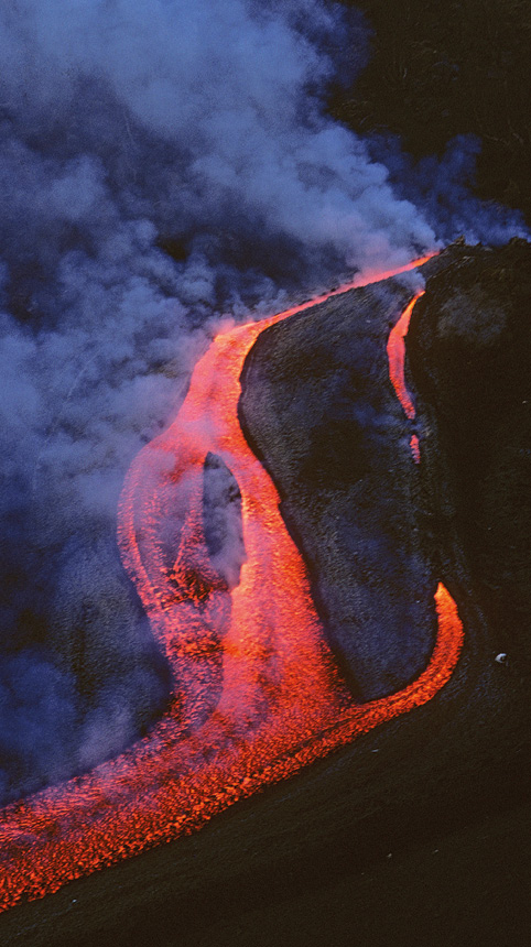 A stream of red liquid lava pours down the side of a blackened slope. Clouds of smoke billow from it.