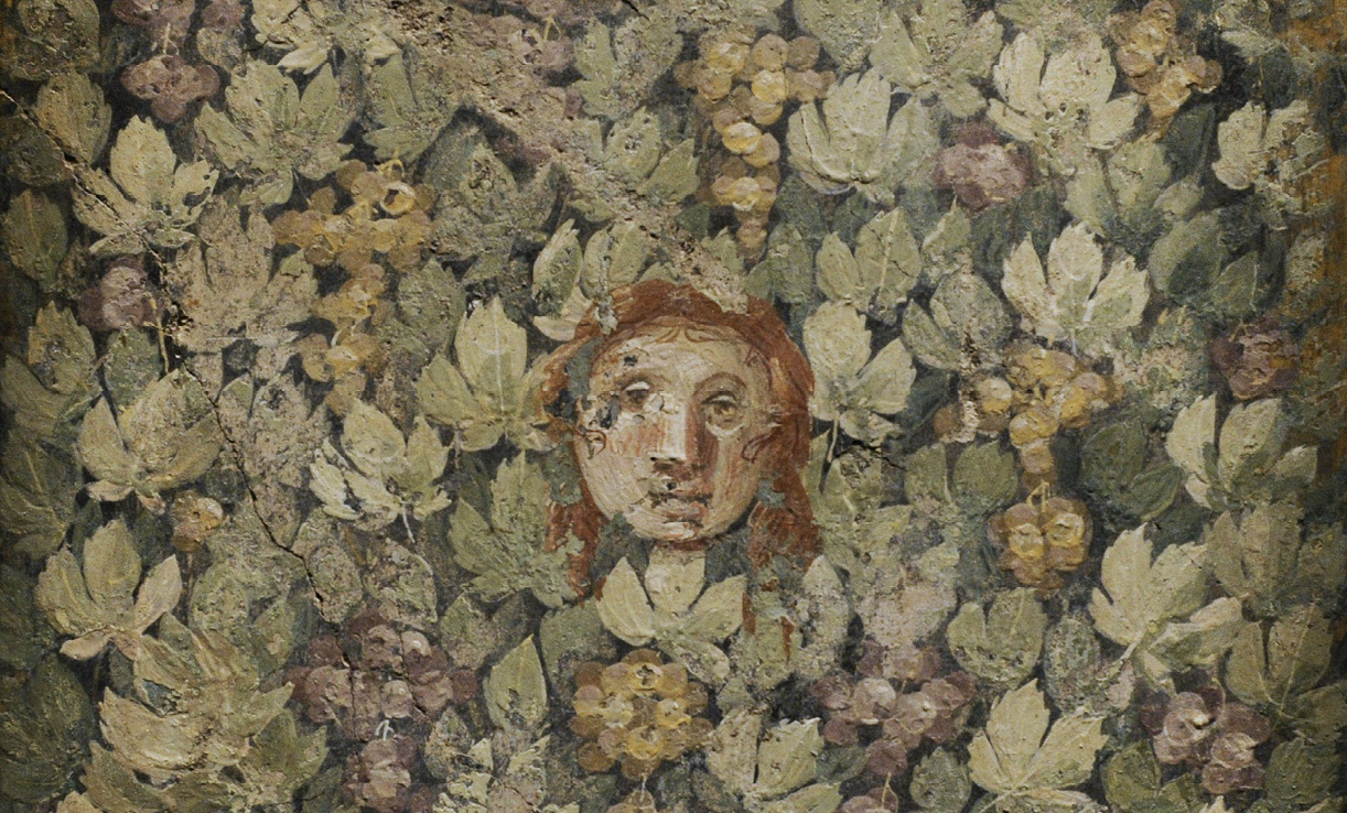 A wall painting showing mask painted with a face and brown hair. It is surrounded by vine leaves of various green colors and bunches of grapes.
