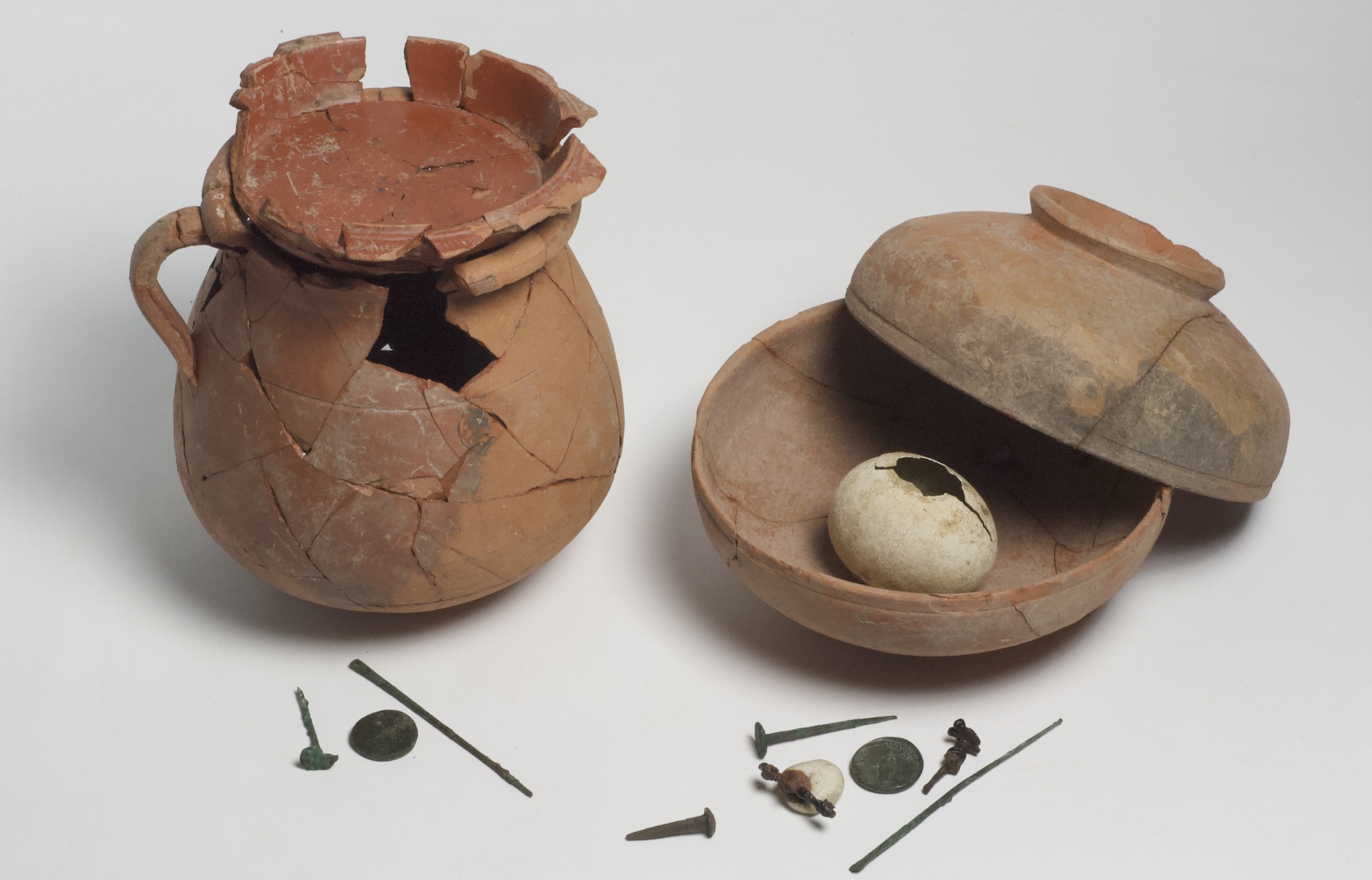 Two terracotta pots with lids. Inside one is an egg. To the side are a few small metal objects including some nails and a coin.