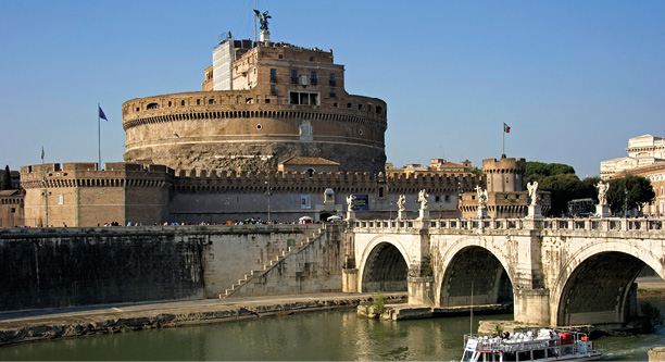 A photo of a circular buidling standing on the edge of a river next to an arched bridge.