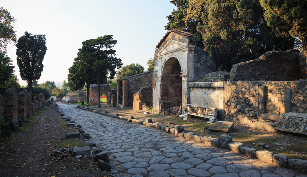 A photo of a Roman paved road with large stone slabs still in place. Along it stand walls and small structures that only partly remain. One has a big arch at the front.