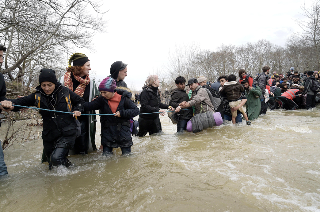 A photo showing a crowd of people carrying bags and wearing winter clothes wading across a river, holding onto a rope line to guide them.