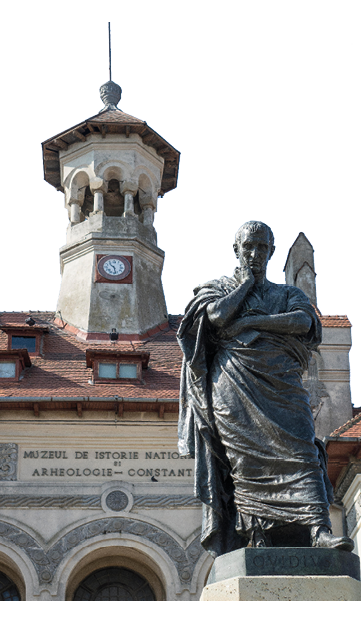 A photo of a statue of a man wearing a toga, looking down and resting his head on his hand. Behind him is a municipal building with a spire.