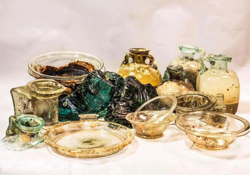 A variety of different glass vessels in different shapes and colors arranged on a pale background. Some are damaged.