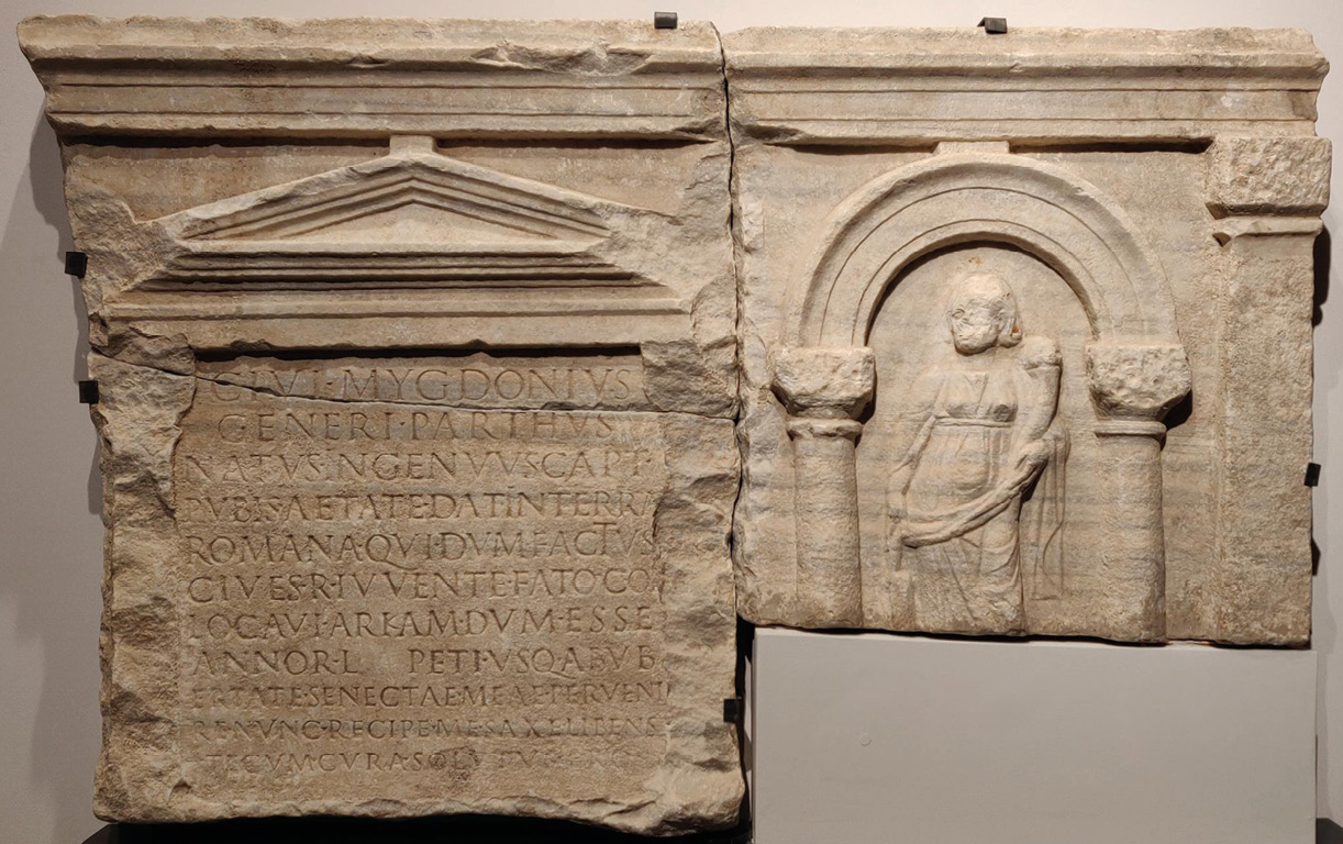 A carved stone relief. On the left is a block of text in captial letters with a pediment above. On the right is an arch under which a female figure stands holding a cornucopia. The relief is chipped and damaged.