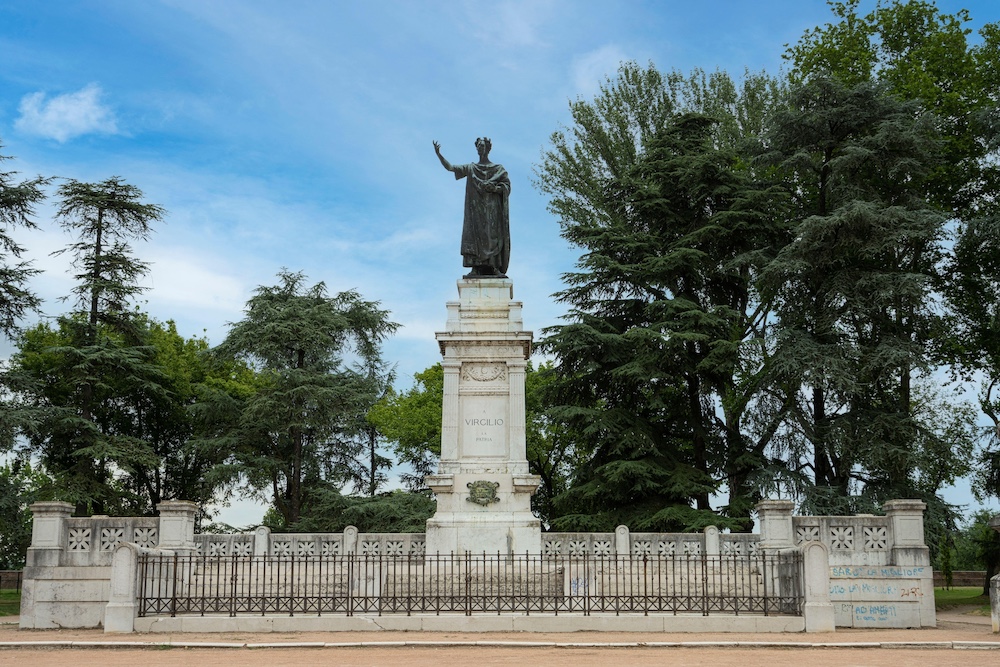 A photo of a modern monument. A white plinth stands in the center on which is a statue of a man with a laurel wreath on this head who is holding his right hand high. Behind are trees and a blue sky.