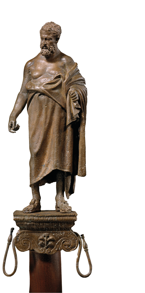 A small metal figure standing on top of a pedestal. He has a beard and a long cloak draped across his left shoulder.