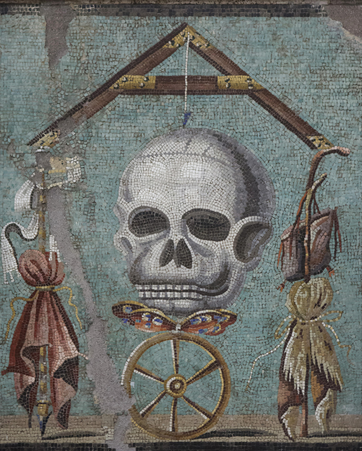 A mosaic with a blue background. In the center is a human skull balanced on a butterfly and and wheel. Hanging on either side is a set of scales.
