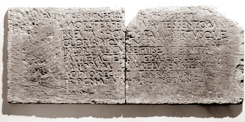 A rectangular stone relief with letters carved on. It is chipped and damaged.