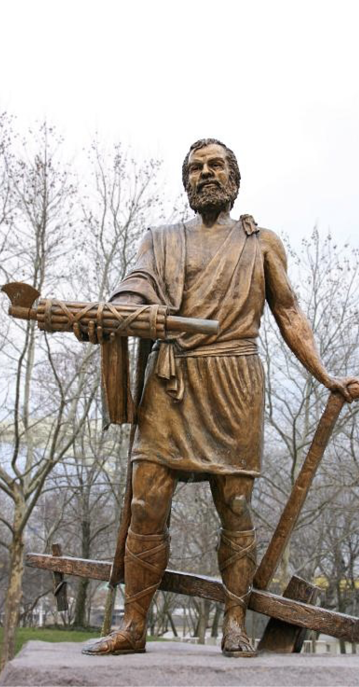 A metal sculpture of a man with a beard. In his outstretched right hand he carries a bundle of sticks. his left hand is behind him holding a plow. He wears a short tunic which ties across one shoulder.