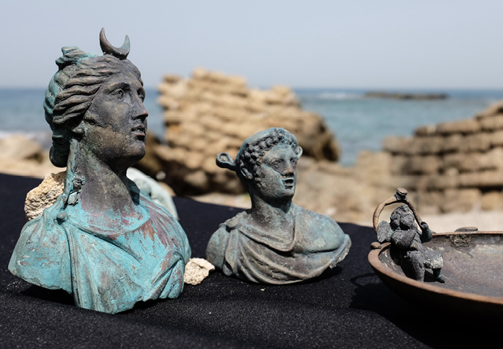 Three small metal statuettes of busts placed on a black piece of material in front of the shore. They are all discolored and sligtly blue. One is a woman with a crescent moon on her head. One is a young man with curly hair, and one is an infant with wings. 