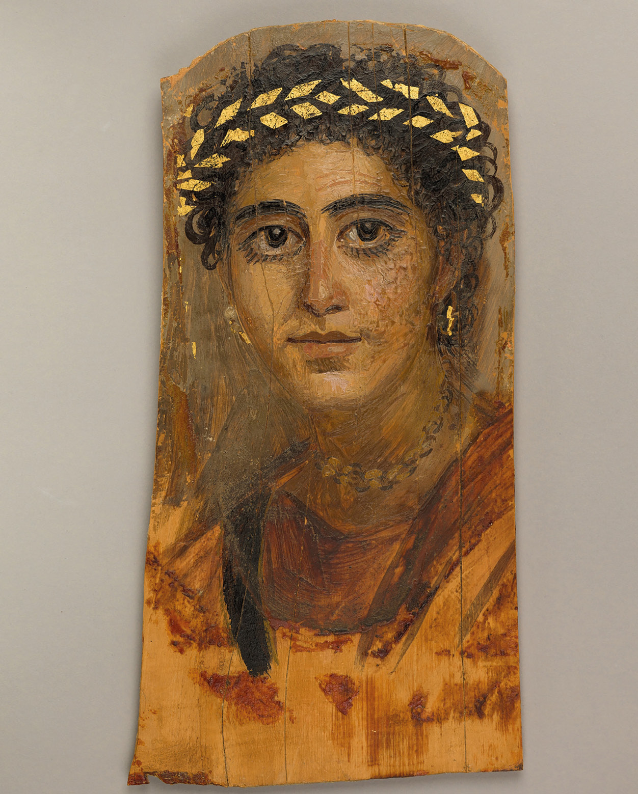 A painted wooden panel showing a woman with dark hair arranged on top of her head. She has thick, dark eyebrows nad long eyelashes, accentuated with makeup. She wears gold and pearl earring and a necklace as well as a gold wreath in her hair. The details of the gold jewelry are picked out with gold leaf.