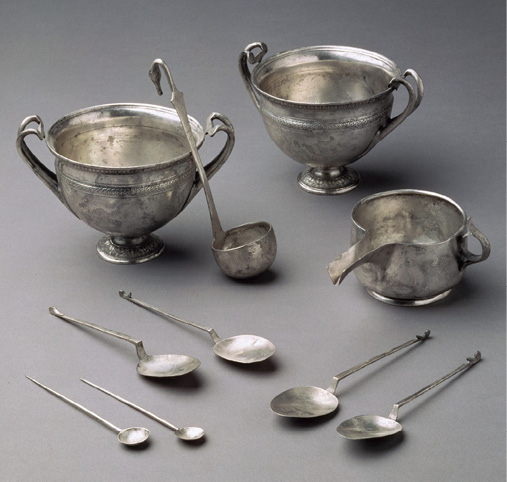A collection of various silver vessels and serving spoons arranged on a grey background.