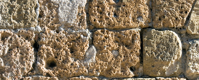 A photo of a wall made of roughly square blocks of rock which have an uneven surface and lots of little holes.