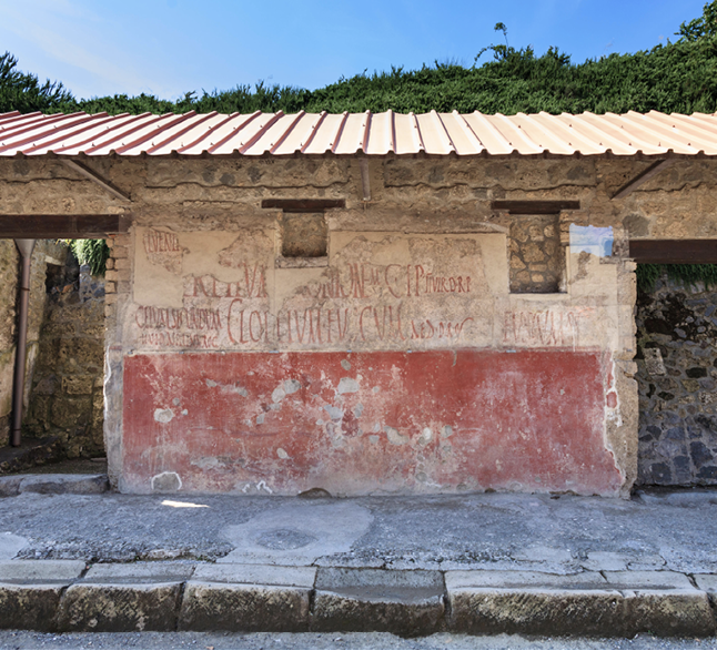 A photo of a wall. The bottom half is painted red and the top half is white with notices in red painted across it. The building has small windows and a modern roof.