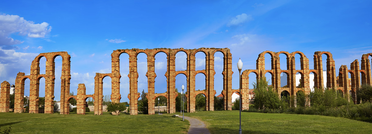 A photo of the remains of a Roman aqueduct standing on a grassy field. At points it is three arches high and made of bricks. Behind is a clear blue sky.