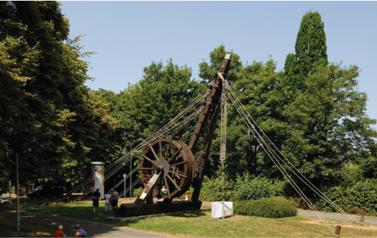 A photo of a large crane with a big wheel and ropes reaching down to the ground. It stands on a patch of grass with trees behind and to the left.