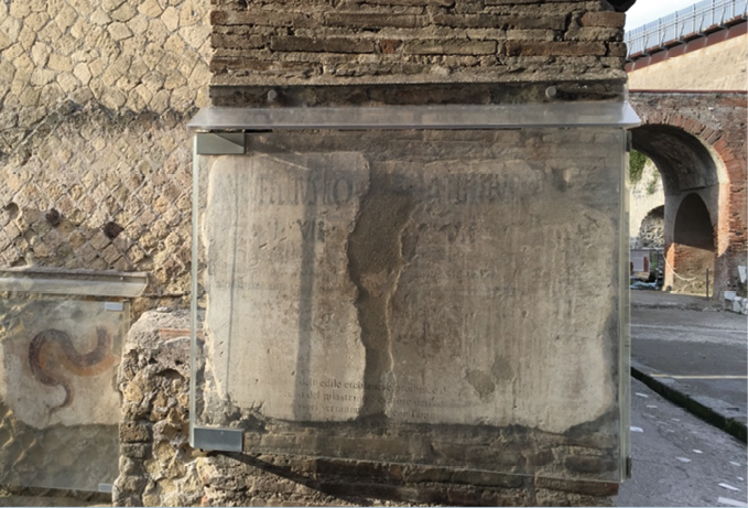 A photo of an brick wall on which a notice is painted. The notice is covered in a sheet of transparent glass. Behind, to the left, is a brick arch.