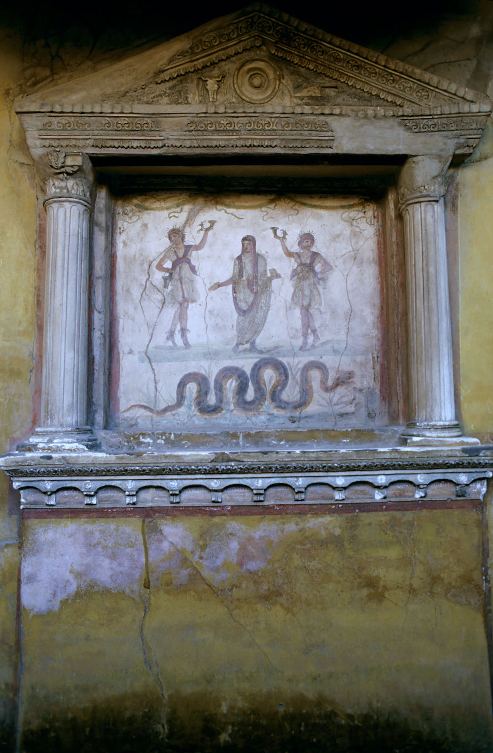 A shrine in the shape of a temple, standing against a wall. Inside is painted a man in a toga with two men on either side with one above their head. Below them is a snake.