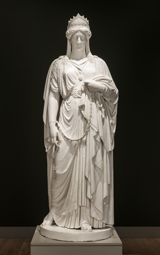 A white stone statue of a woman against a black background. The woman has a long dress, gathered under the breast, and wears a crown. Her hands are tied together with a chain which hangs between them.
