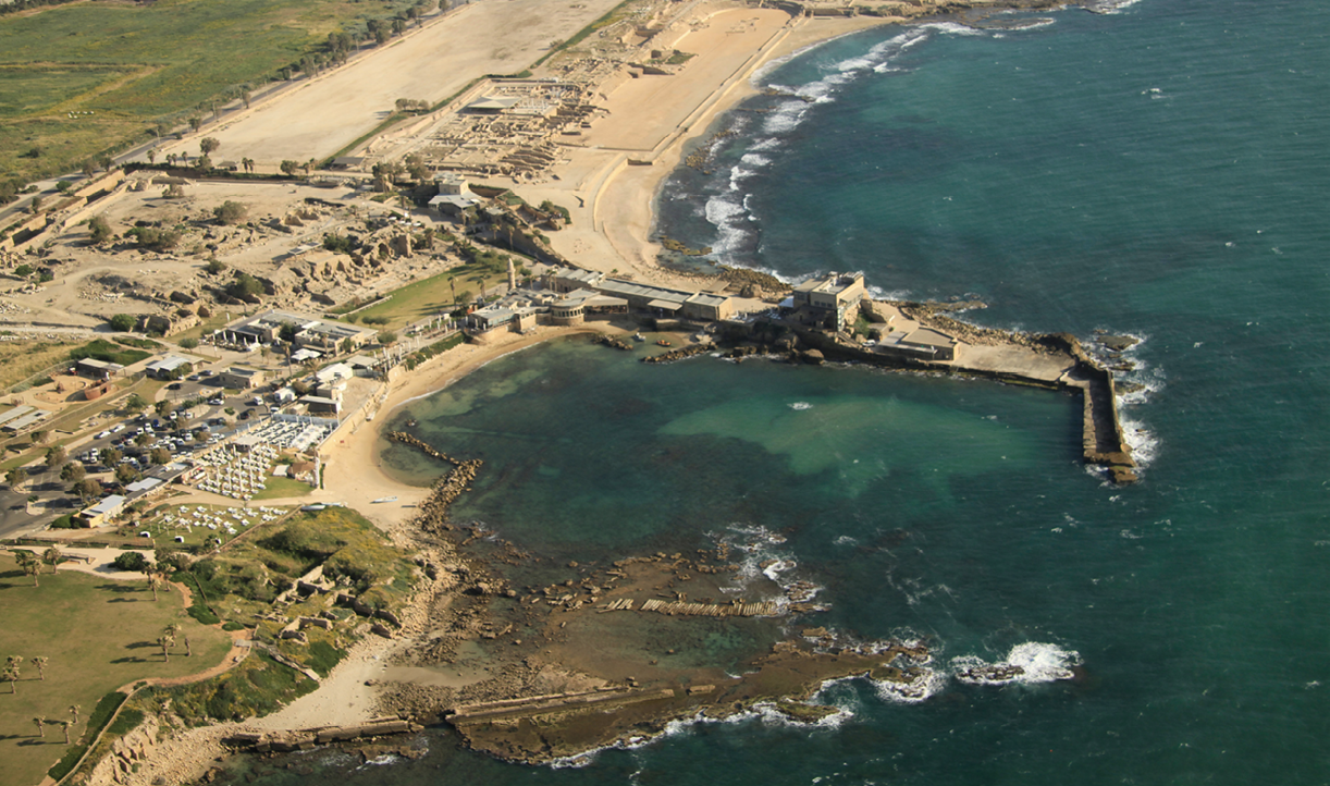 A photo of a shoreline from above. Reaching out into the sea is a harbor. On the shore is a sandy beach, a grassy area and low buildings.