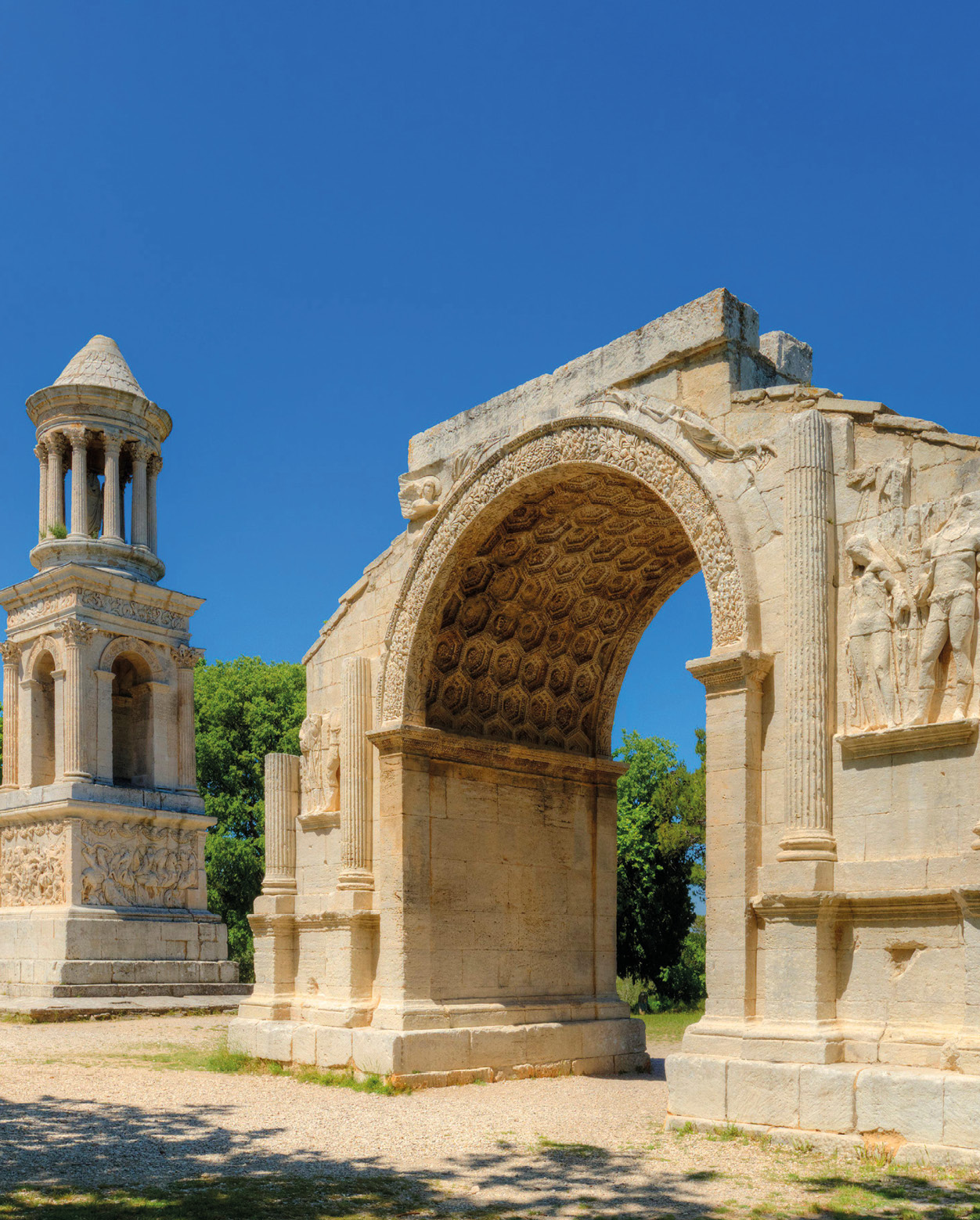 A photo of a stone arch with pillars carved into the side and figures. The underside of the arch is carved with a hexagonal pattern. Behind the arch is another stone monument with a circular top.