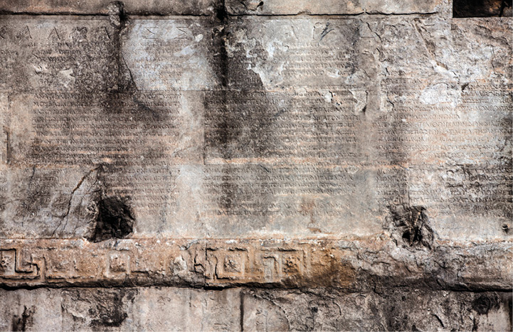 A stone wall on which is inscribed rows of text. It is chipped and damaged. A geometric pattern is just visible along the bottom.
