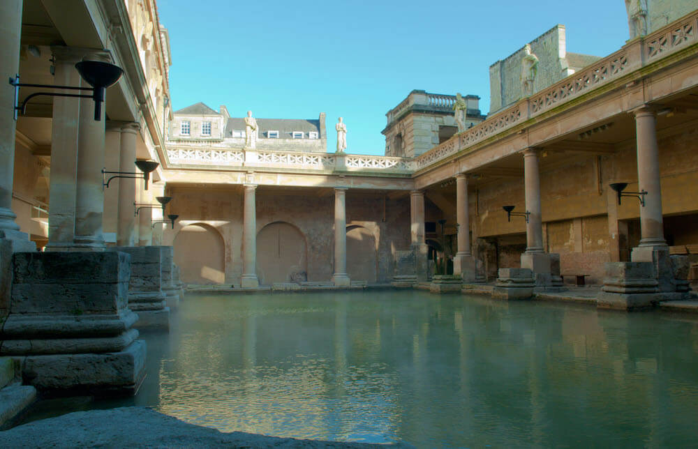 A photograph of the Great Bath at Aquae Sulis. The bath is surrounded by a covered walkway and has no roof.