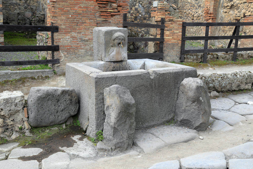A large public fountain made of grey stone. A face is carved into one part; water would have emerged from its mouth.