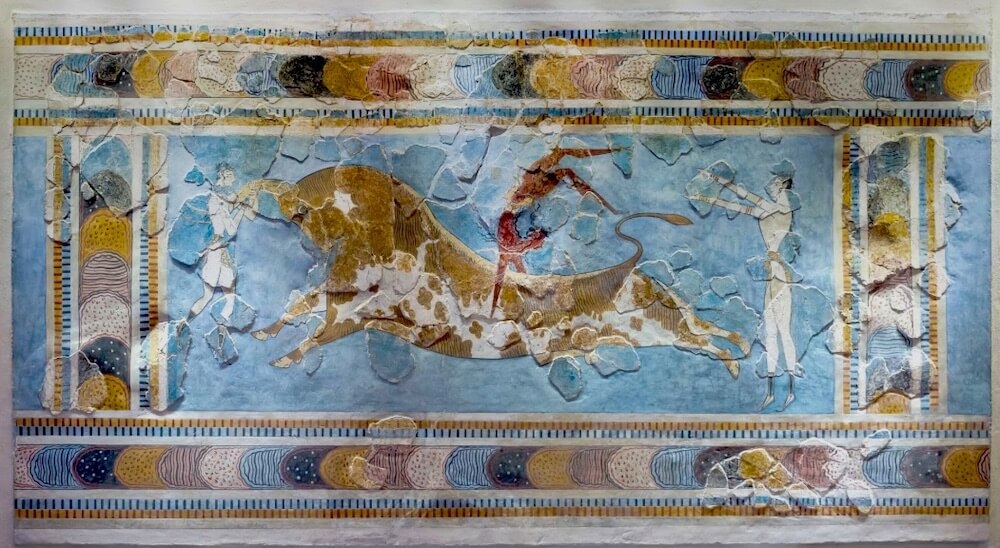 Fresco (painting on plaster) showing a man leaping over a bull, with his legs in the air and his hands on its flanks. Two people watch, one on either side of the bull. The background is a faded blue, and the border is decorated with overlapping coloured circles and a checkerboard pattern.