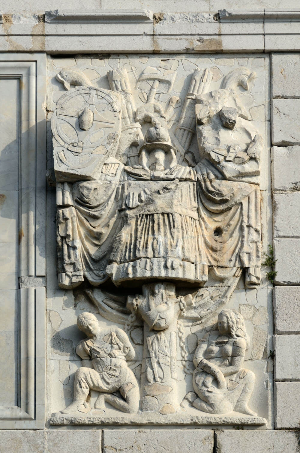 A stone carving from a triumphal monument showing two enslaved captives, one male and one female, chained at the bottom of a Roman trophy.