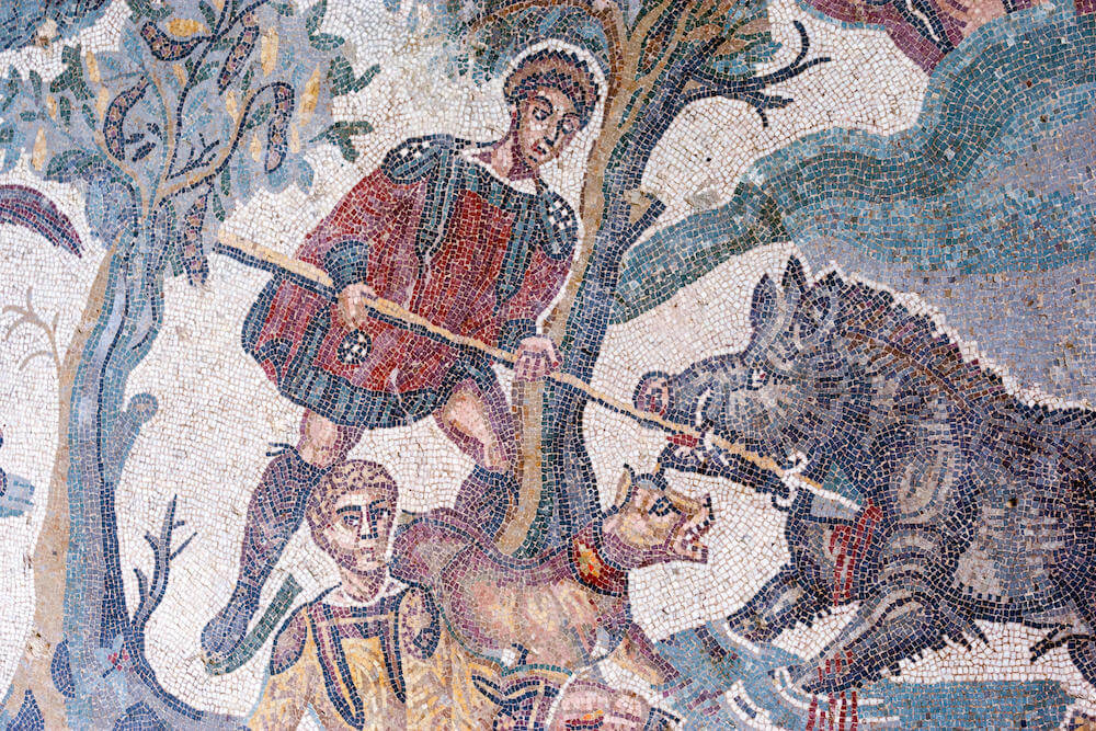 Mosaic showing a man in a tunic and cape fighting off a large boar with a spear. At his feet a hinting dog is growling at the boar. Another man lies nearby.
