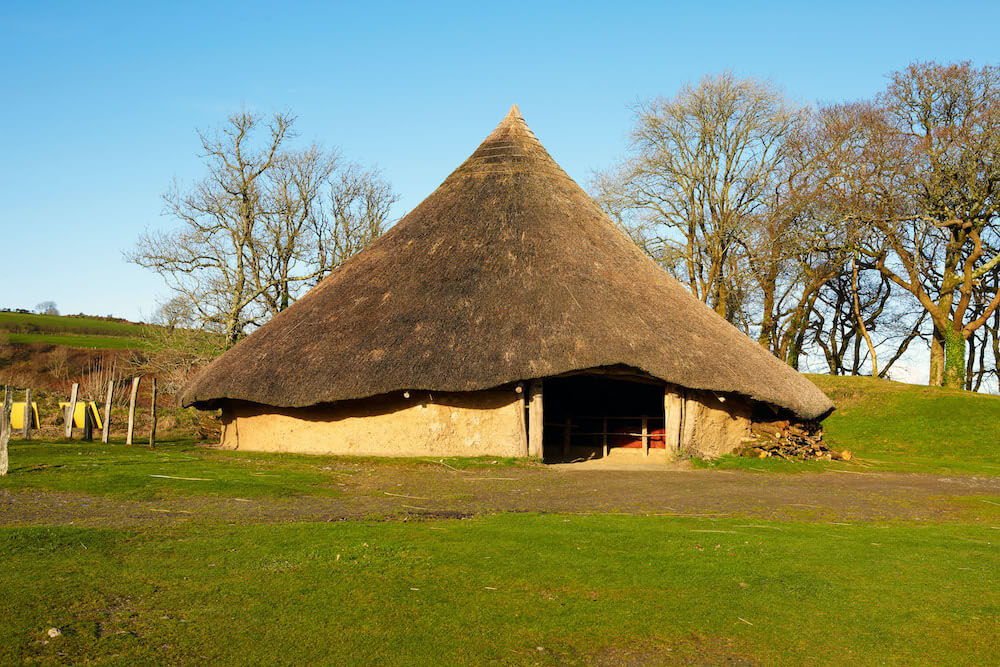 An image of a replica Celtic roundhouse with low wattle-and-daub walls and a high, pointed straw roof.