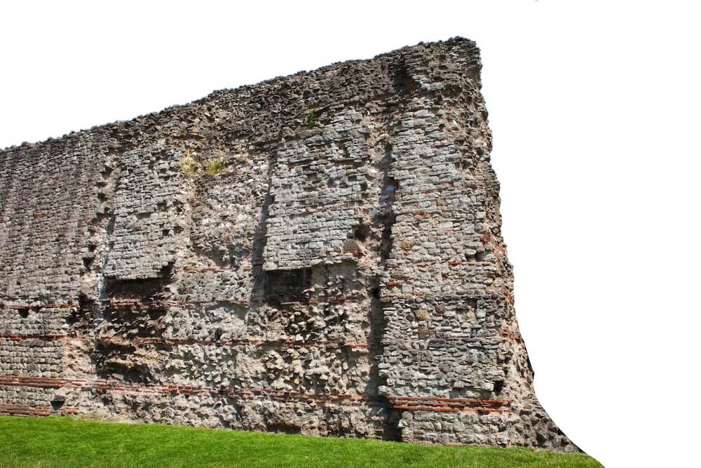 An image of a stone wall containing layers of brick. Although mossy with a few stones having fallen out, the wall still stands tall.
